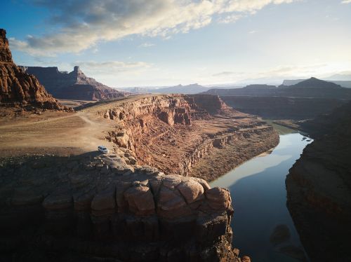 Scenic view of a river flowing through a canyon landscape with a vehicle on a cliffside road, under a blue sky with scattered clouds.