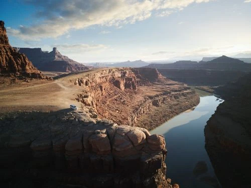 Scenic view of a river flowing through a canyon landscape with a vehicle on a cliffside road, under a blue sky with scattered clouds.
