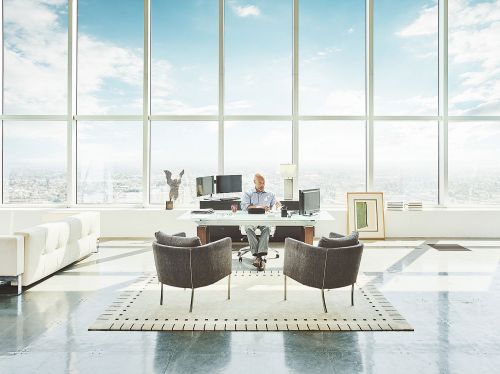 Man in a modern office with expansive windows, sitting at a desk with computer monitors, facing the city skyline and blue sky.