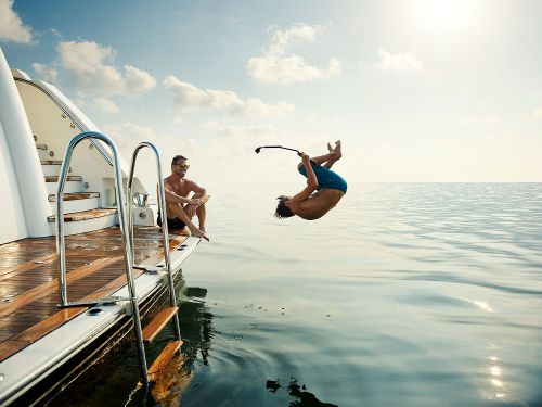 Two men enjoy a sunny day on a yacht, one performs a backflip into the sea, while the other relaxes on the edge of the boat.