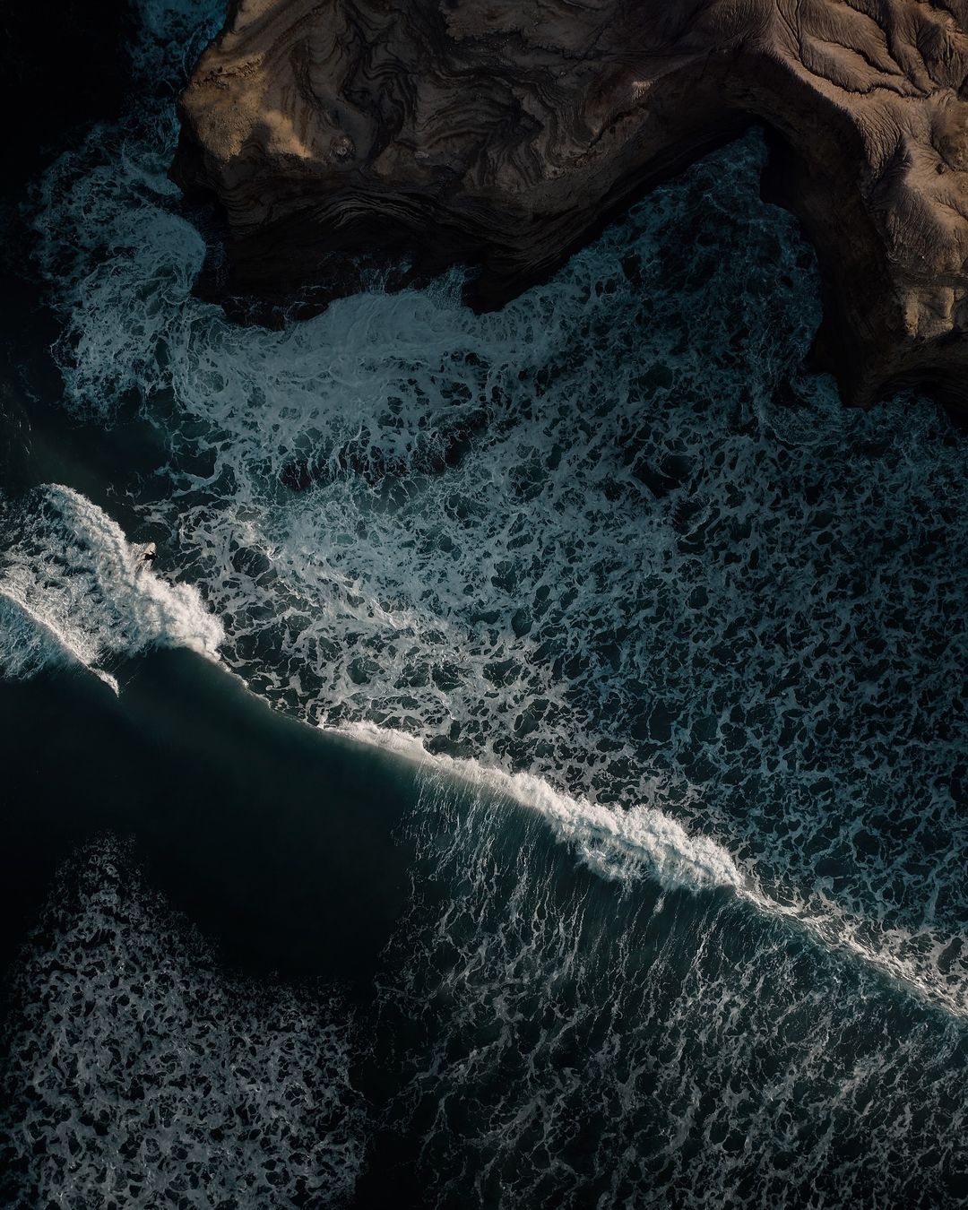 Aerial view of ocean waves crashing against a dark sand beach on the coastline, creating white foam and contrasting with the blue water.