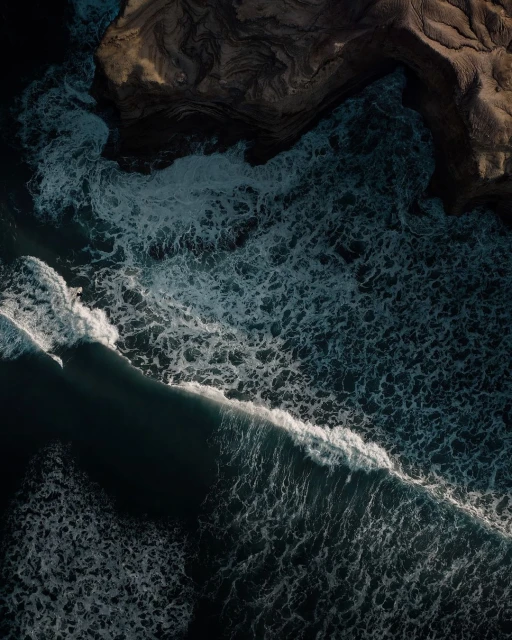 Aerial view of ocean waves crashing against a dark sand beach on the coastline, creating white foam and contrasting with the blue water.