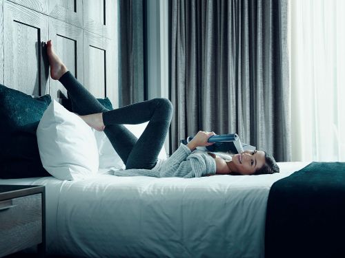 A woman with long hair is lying on a bed in a bedroom reading a magazine, with her feet resting against the headboard.