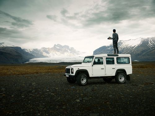 Adult male photographer standing on a white SUV in Iceland with snow-capped mountains, gravel ground, and cloudy skies in the background.