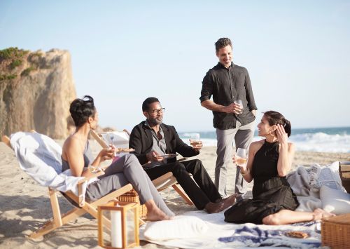 Four adults relax on a beach with a rock formation and ocean. One man stands holding a wine glass while the others sit and socialize.