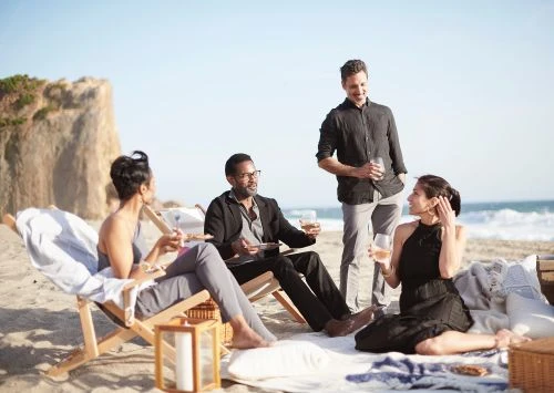 Four adults relax on a beach with a rock formation and ocean. One man stands holding a wine glass while the others sit and socialize.