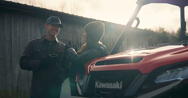 Couple leaning on a red Kawasaki UTV outdoors, wearing caps and jackets on a sunny day, enjoying a drink outside with a wooden fence.