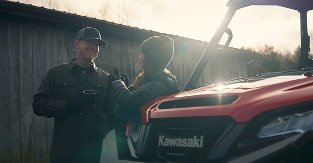 Couple leaning on a red Kawasaki UTV outdoors, wearing caps and jackets on a sunny day, enjoying a drink outside with a wooden fence.