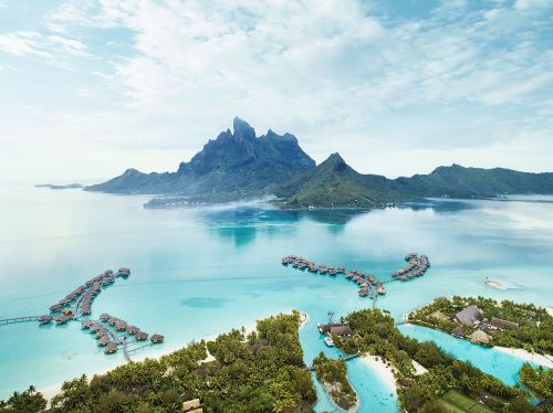 Aerial view of luxury overwater bungalows and villas at tropical island resort near mountainous coastline in turquoise water.