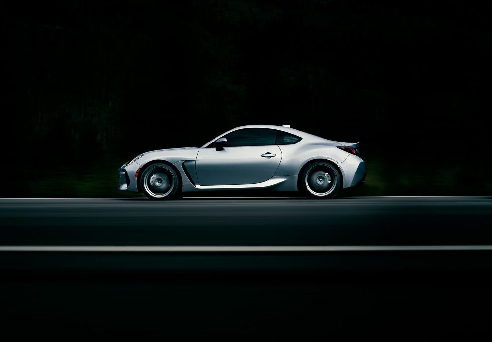 Side view of a silver sports car on a dark asphalt road, blurred background with hints of greenery, showcasing sleek automotive design.