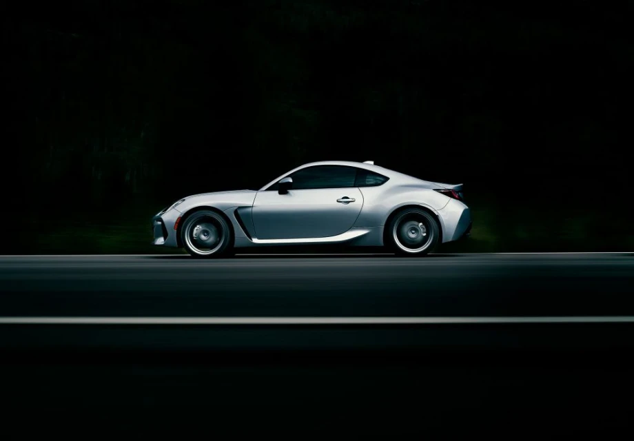 Side view of a silver sports car on a dark asphalt road, blurred background with hints of greenery, showcasing sleek automotive design.