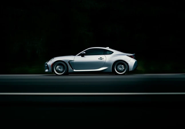 Side view of a silver sports car on a dark asphalt road, blurred background with hints of greenery, showcasing sleek automotive design.