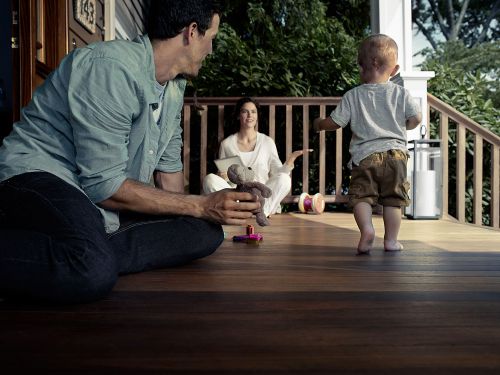 Family scene on a porch: father holding a stuffed animal, toddler walking barefoot, and mother sitting in the background.
