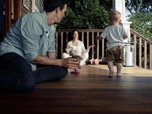 Family scene on a porch: father holding a stuffed animal, toddler walking barefoot, and mother sitting in the background.