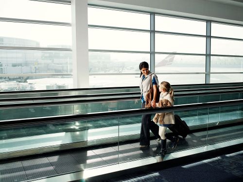 Mother and daughter walking on a moving walkway in an airport, pulling a suitcase. Large windows and airplanes in the background.