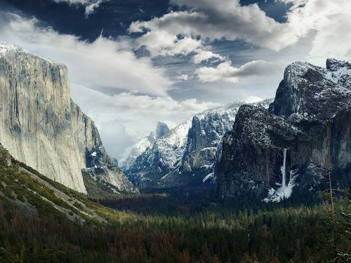 Scenic view of Yosemite Valley with snowy mountains, evergreen forest, and dramatic clouds, showcasing nature's grandeur and tranquil beauty.