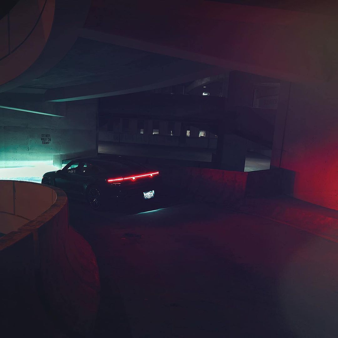 A dark-colored car is parked under a concrete overpass at night with red and teal lighting illuminating the dark surroundings.