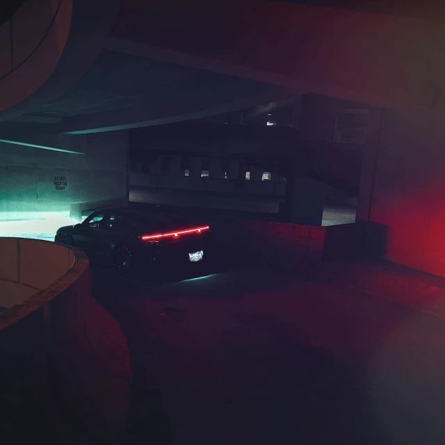 A dark-colored car is parked under a concrete overpass at night with red and teal lighting illuminating the dark surroundings.