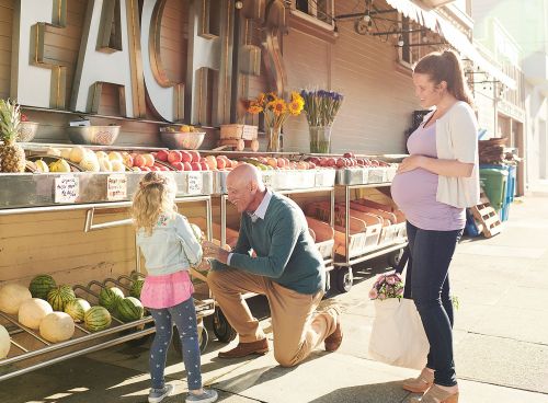 Three people shop at an outdoor fruit and vegetable market; a pregnant woman, an older man and a young girl near produce.