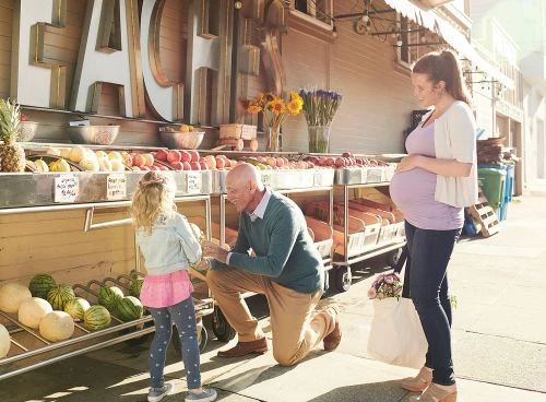 Three people shop at an outdoor fruit and vegetable market; a pregnant woman, an older man and a young girl near produce.