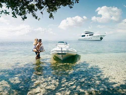 Couple standing in ocean water next to boat and yacht with a sunny sky and puffy clouds in the background; vacation lifestyle image.