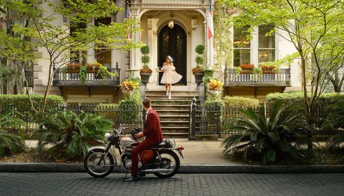 Retro-style scene with a woman in a polka dot dress descending steps of a classic building toward a man on a vintage motorcycle.