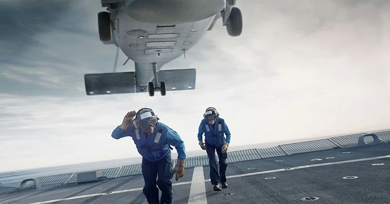 Two Navy sailors in blue uniforms run on an aircraft carrier flight deck under a landing helicopter, signaling its approach on a cloudy day.