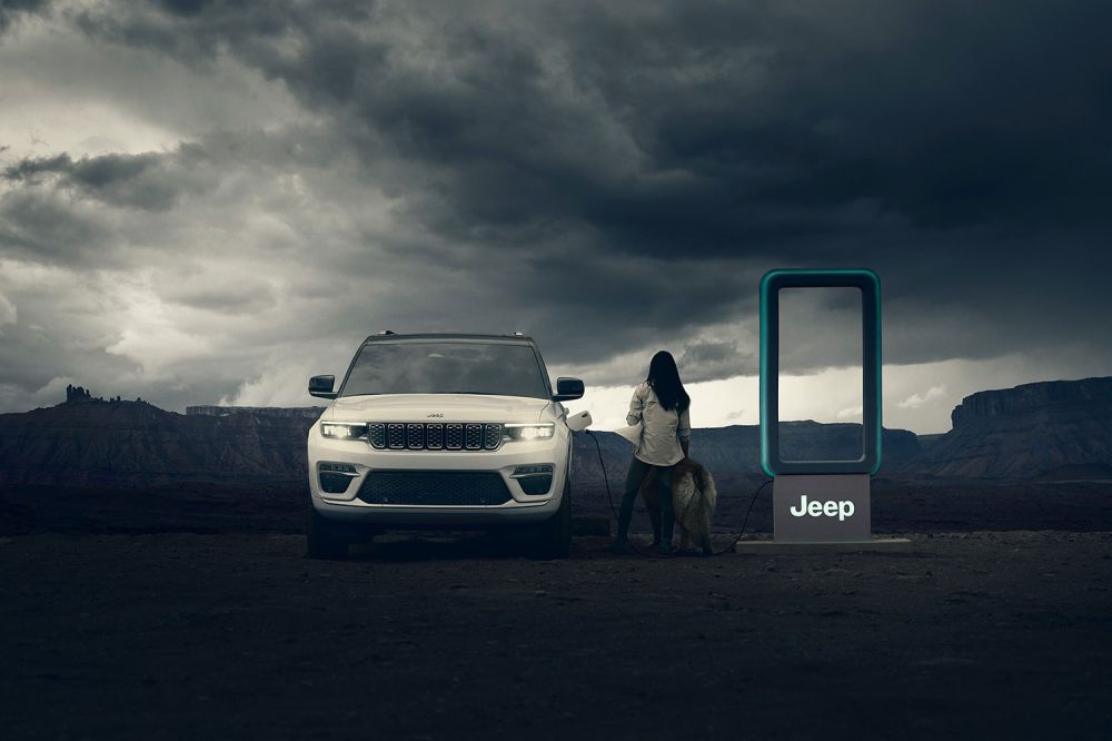 A white Jeep Grand Cherokee 4xe charges at a station in the desert with a woman and dog, set against a cloudy sky.