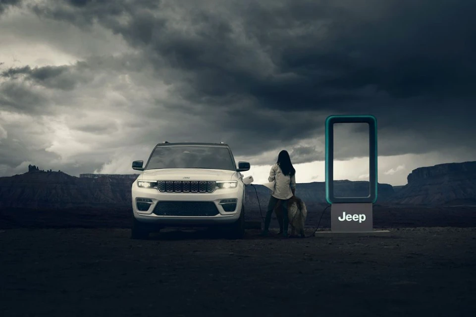 A white Jeep Grand Cherokee 4xe charges at a station in the desert with a woman and dog, set against a cloudy sky.