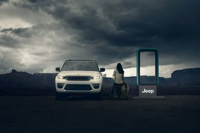 A white Jeep Grand Cherokee 4xe charges at a station in the desert with a woman and dog, set against a cloudy sky.