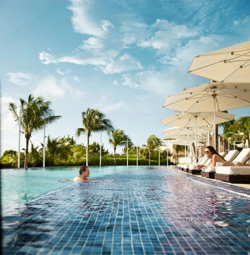 Outdoor pool scene with adults swimming, sun loungers under parasols, and palm trees against a bright sky. Summer vacation vibe.