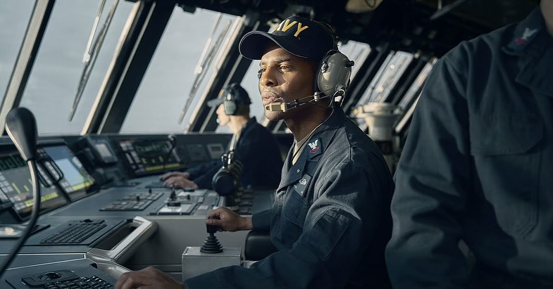 Sailor in navy uniform operates ship controls, with view of open water through windows, nautical scene in muted blue and gray tones.