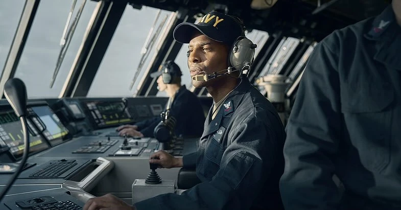 Sailor in navy uniform operates ship controls, with view of open water through windows, nautical scene in muted blue and gray tones.