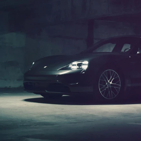 Low-angle shot of a dark gray Porsche Taycan in an indoor garage, featuring its illuminated headlights and sleek design.