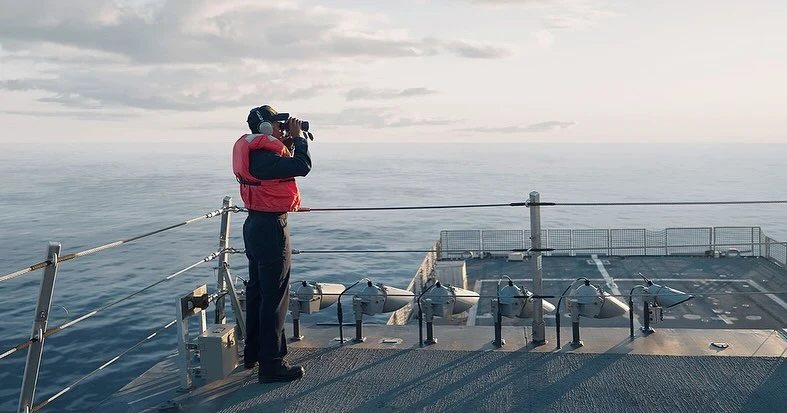 Man in life vest looks through binoculars on ship deck over ocean, under soft cloudy sky, providing a naval perspective.