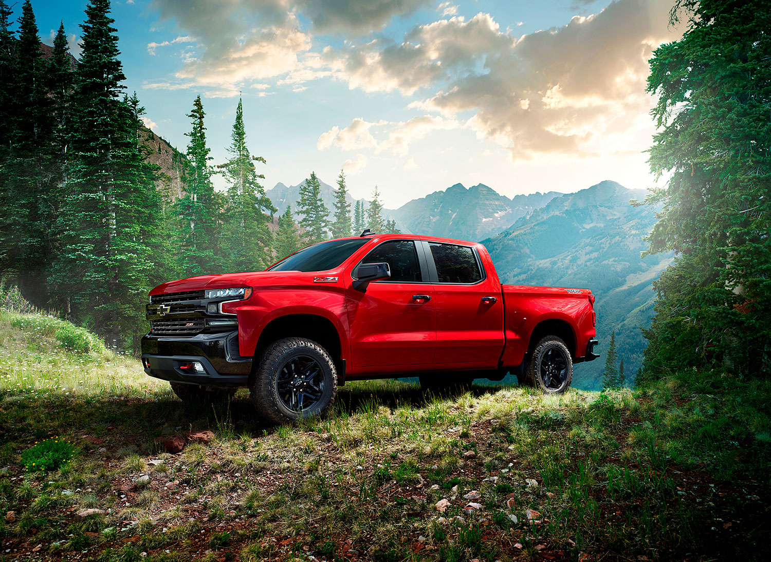 Red Chevrolet Silverado pickup truck is parked on a grassy hill in a mountainous forest landscape under a sky with clouds.