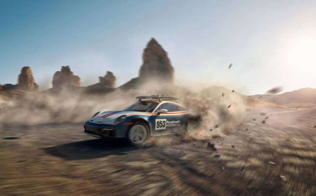 A blue and white Porsche rally car speeds through a desert landscape, kicking up dust and rocks, set against rock formations and a clear sky.