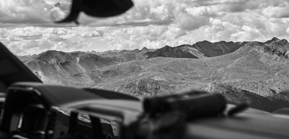 Monochrome mountain landscape view from inside a vehicle, dramatic sky, and foreground automobile elements.