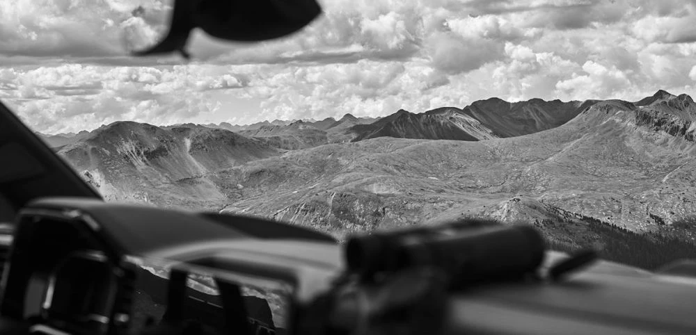 Monochrome mountain landscape view from inside a vehicle, dramatic sky, and foreground automobile elements.