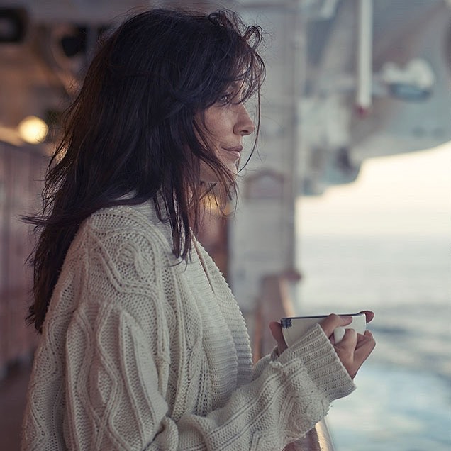 Adult woman with dark hair in white cable-knit cardigan holds a cup indoors on a ship or boat, looking out towards the ocean.