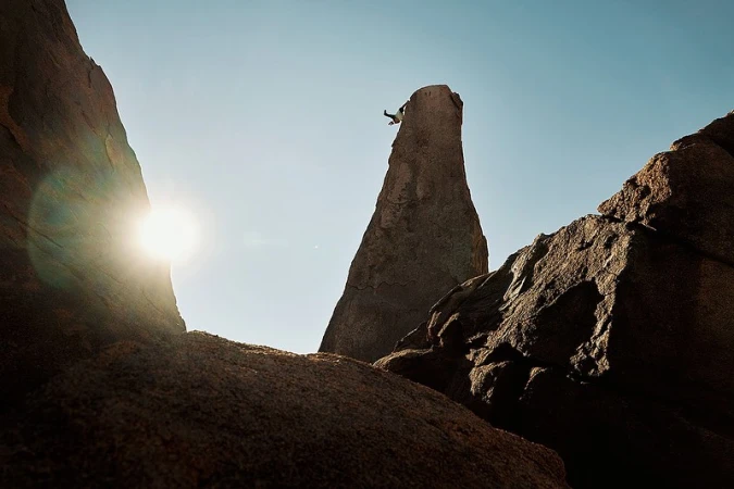 Rock climber ascending a tall, slender rock formation under a clear sky, framed by larger rock structures and sunlight on a sunny day.