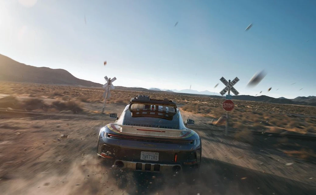 Rear view of a Porsche sports car speeding down a dirt road in a desert landscape with railroad crossing signs and clear blue sky.