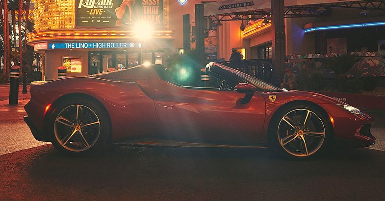 Red Ferrari convertible parked on a city street at night, with bright lights from nearby buildings illuminating the scene.