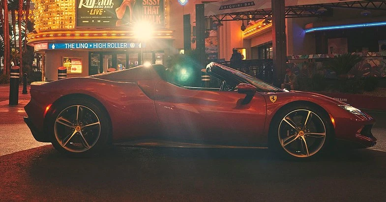 Red Ferrari convertible parked on a city street at night, with bright lights from nearby buildings illuminating the scene.