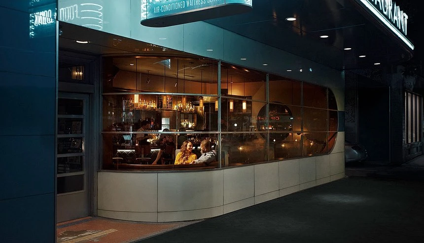 Exterior shot of a restaurant at night with a couple sitting near the window and the restaurant name in lights above the door.