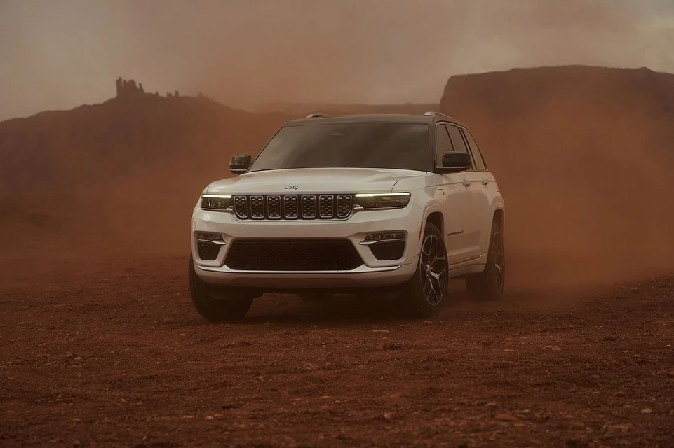 A white Jeep Grand Cherokee SUV stands in a desert environment with distant rock formations and blowing sand, showcasing automotive design.