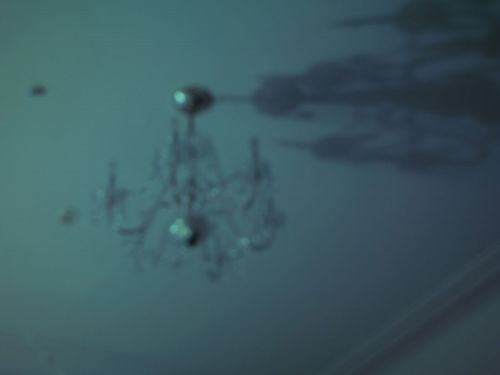 Underexposed shot of a crystal chandelier hanging from the ceiling of a dimly lit room, giving a vintage or haunted-house impression.