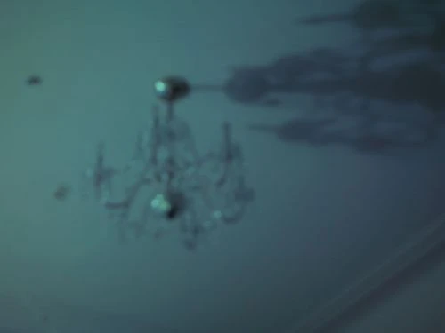 Underexposed shot of a crystal chandelier hanging from the ceiling of a dimly lit room, giving a vintage or haunted-house impression.