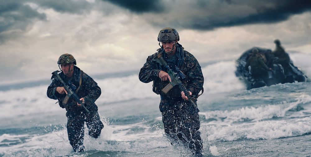 Two military personnel in camouflage gear advancing from the ocean towards the shore, with a dark sky and an inflatable boat in the distance.