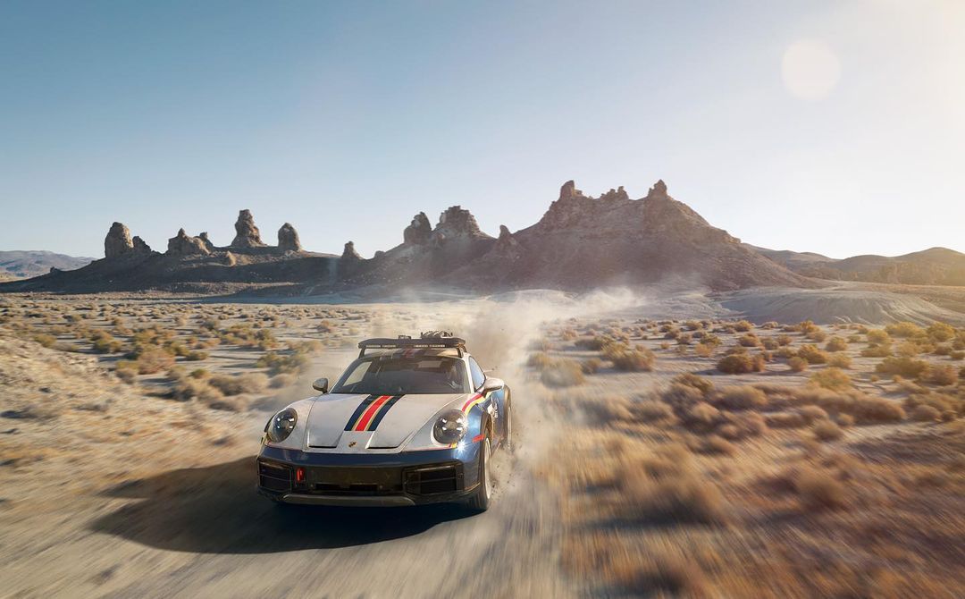 A Porsche 911 Dakar speeds across a desert landscape, kicking up dust with rocky mountains and a bright blue sky in the background.
